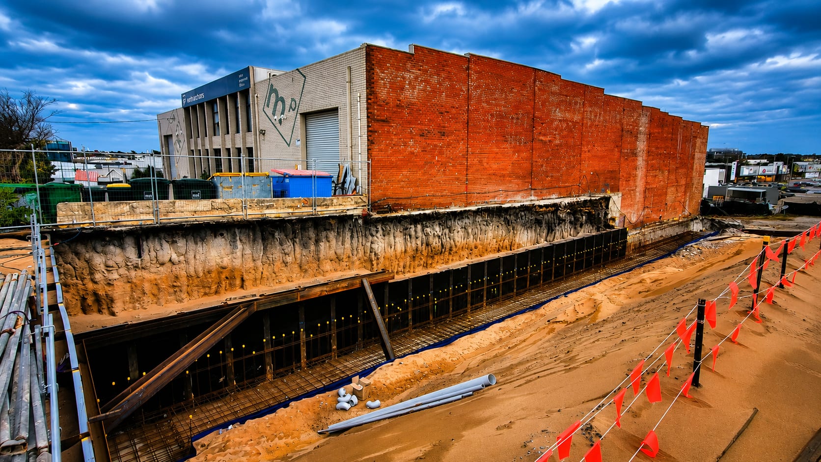Construction scene showing a red-brick building above an excavation with exposed ground, formwork and reinforcing steel